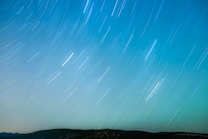 Long exposure photograph capturing the trails of stars moving across the night sky, with a landscape of dark hills at the bottom.