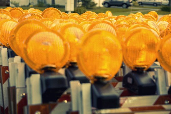 A large group of bright orange construction safety lights arranged closely together, giving a sense of repetition and uniformity. The lights are attached to barricades, typically used in traffic management.