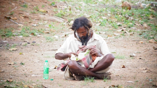 A man with unkempt hair and a beard sits on the ground outdoors with a bottle of green liquid next to him. He is focused on something in his hands, which is wrapped in a cloth. The surroundings are natural with dry grass and scattered leaves, giving a rustic and rural feel.
