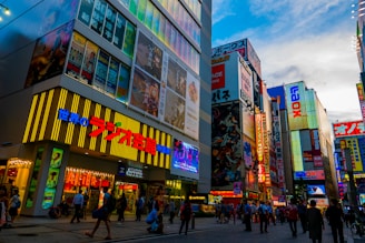 A vibrant urban street scene featuring tall buildings covered in colorful advertisements and billboards, including various Asian characters and graphics. The street is bustling with people walking, and the sky above is partly cloudy.