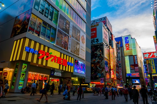 A vibrant urban street scene featuring tall buildings covered in colorful advertisements and billboards, including various Asian characters and graphics. The street is bustling with people walking, and the sky above is partly cloudy.