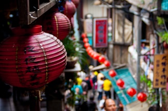 Red lanterns with Chinese characters hang prominently in a bustling street market setting. The background features a crowd of people, some blurred, moving through the narrow passage lined with shops. Vibrant signage and greenery add to the lively atmosphere.