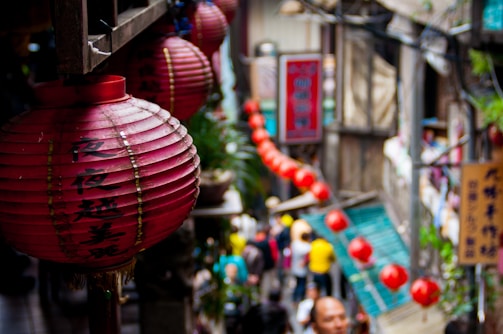Red lanterns with Chinese characters hang prominently in a bustling street market setting. The background features a crowd of people, some blurred, moving through the narrow passage lined with shops. Vibrant signage and greenery add to the lively atmosphere.