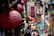 Red lanterns with Chinese characters hang prominently in a bustling street market setting. The background features a crowd of people, some blurred, moving through the narrow passage lined with shops. Vibrant signage and greenery add to the lively atmosphere.