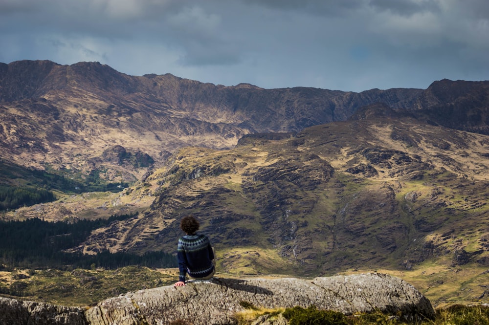 person sitting on concrete barrier overlooking mountain at daytime