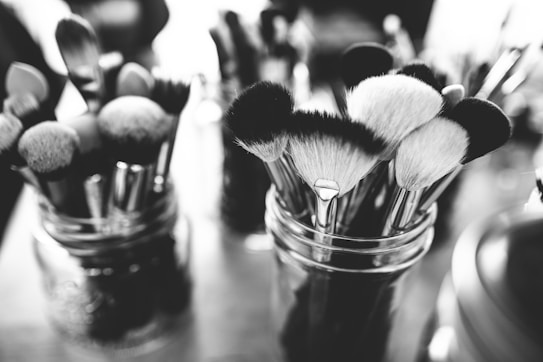 A collection of makeup brushes of various sizes and shapes is placed inside glass jars. The image is in black and white, giving it a classic and timeless feel. The background is blurred, accentuating the focus on the makeup brushes.
