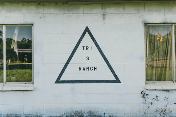 A white building facade with two windows, each featuring weathered curtains. The wall displays a black-outlined triangle with the words 'TRI S RANCH' inside it. Greenery is visible through the windows, adding a natural element.