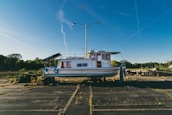 A boat is positioned on land in a deserted parking lot with overgrown grass. It has a white and blue color scheme and is situated beneath a tall streetlight. The background features a clear blue sky with visible contrails and some greenery on the periphery.