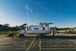 A boat is positioned on land in a deserted parking lot with overgrown grass. It has a white and blue color scheme and is situated beneath a tall streetlight. The background features a clear blue sky with visible contrails and some greenery on the periphery.
