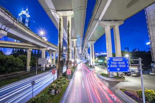 A dynamic urban scene featuring elevated highways and overpasses crisscrossing against a vibrant blue evening sky. Bright lights and light trails from vehicles create a sense of motion and speed, while skyscrapers and trees are visible in the background. Signs and road markings add to the complex infrastructure of the city.