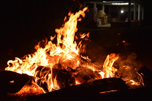 A large, intense fire burns brightly, with tall flames and glowing embers visible in the foreground. The background is dimly lit, featuring a wooden structure with a roof, possibly a gazebo or outdoor shelter.