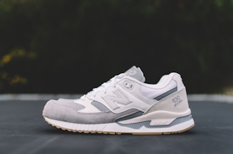 A white and gray athletic shoe with a modern design is positioned on a flat surface against a blurred dark background. The shoe features a mix of materials, including mesh and suede, and showcases detailed stitching with a visible logo and embossed text on the side.