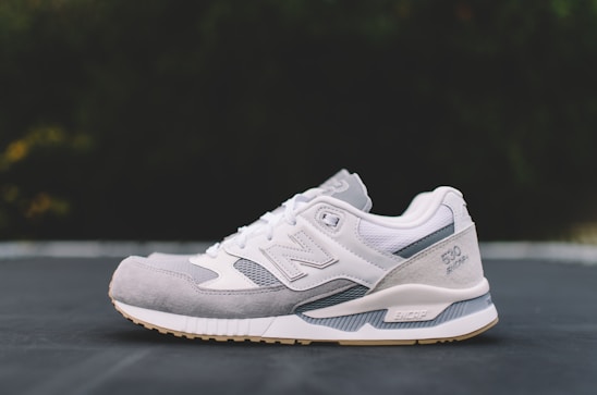 A white and gray athletic shoe with a modern design is positioned on a flat surface against a blurred dark background. The shoe features a mix of materials, including mesh and suede, and showcases detailed stitching with a visible logo and embossed text on the side.