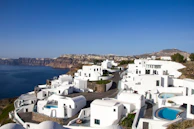 Whitewashed buildings with bright blue domes and swimming pools are perched along a clifftop, overlooking a deep blue sea. In the background, rugged cliffs extend into the distance under a clear blue sky.