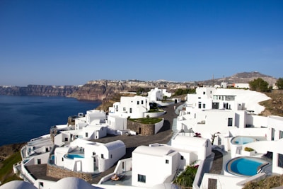 Whitewashed buildings with bright blue domes and swimming pools are perched along a clifftop, overlooking a deep blue sea. In the background, rugged cliffs extend into the distance under a clear blue sky.