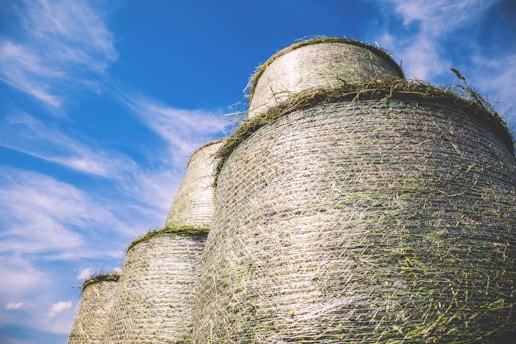 Large cylindrical hay bales stacked against a bright blue sky, with wispy white clouds creating a serene backdrop. The bales are wrapped in mesh, giving them a textured appearance, and they catch the sunlight, highlighting their natural color.