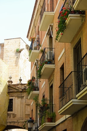 A row of apartment buildings with several balconies visible, adorned with green plants and red flowers. The architecture features brick walls, wrought iron railings, and narrow windows. There is an older stone building in the background, partially covered by a yellow tarp.