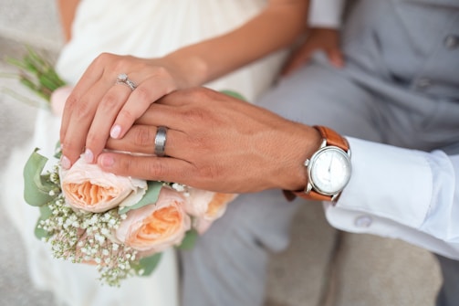 Close-up of their intertwined hands with wedding rings, resting on a bouquet of wildflowers.
