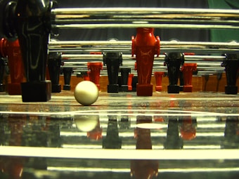 A close-up view from the field level of a foosball table, featuring red and black foosball player figures lined up on metal rods. A white foosball ball is positioned in the foreground on the reflective surface of the table, with its reflection clearly visible. The perspective creates a dynamic and somewhat tense atmosphere, reminiscent of a paused moment in a game.