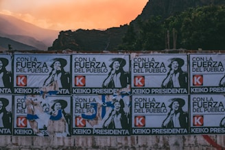 A series of political posters are pasted on a wall, set against a backdrop of mountainous landscape and a warm, orange-tinted sky. The posters feature a person in a hat, with the text 'Con la Fuerza del Pueblo' and 'Keiko Presidenta' prominently displayed, along with a red 'K' logo.