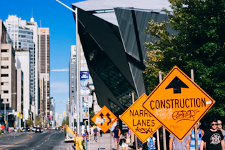 Several orange construction signs are positioned along a busy city street with high-rise buildings and a modern, angular architectural structure. People are walking on the sidewalk alongside the signs, and a few cars can be seen on the road. Trees and greenery are visible, adding a touch of nature to the urban setting.
