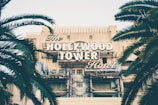 A vintage-style sign displaying 'The Hollywood Tower Hotel' is prominently featured against the backdrop of an old, slightly worn building. Palm trees frame the composition on both sides, adding a touch of greenery to the scene.