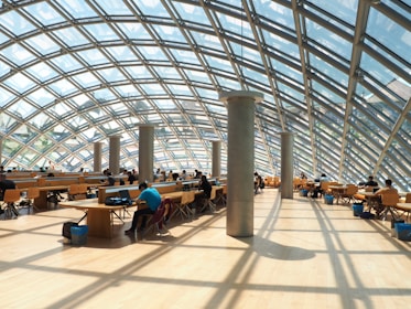 A spacious interior with a modern glass and steel dome structure allowing natural light to fill the space. Rows of desks and chairs are occupied by people engaged in work or study. The wooden floor and minimalist design create a warm and inviting atmosphere.