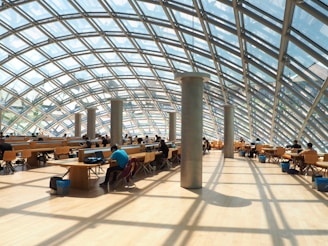 A spacious interior with a modern glass and steel dome structure allowing natural light to fill the space. Rows of desks and chairs are occupied by people engaged in work or study. The wooden floor and minimalist design create a warm and inviting atmosphere.