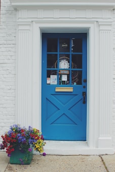 A bright blue door with a traditional design is set within a white brick wall, featuring a golden mail slot and black handle. Various notices and a 'closed' sign are displayed in the door's window panes. Vibrantly colored flowers in a green pot sit to the left of the entrance, enhancing the welcoming appearance.