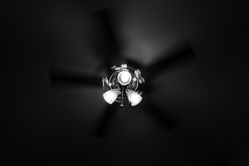 A ceiling fan is captured from below, with three light fixtures attached. The fan blades are blurred, suggesting motion, while the central light illuminates the surrounding area.