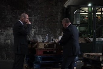 Two men in suits are standing near a stack of wooden pallets in an industrial setting. One man appears to be eating from a container while the other stands nearby, possibly engaged in conversation. The background includes a brick wall and industrial equipment.