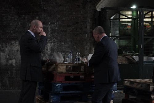 Two men in suits are standing near a stack of wooden pallets in an industrial setting. One man appears to be eating from a container while the other stands nearby, possibly engaged in conversation. The background includes a brick wall and industrial equipment.