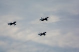 Three military jet aircraft are flying in formation against a cloudy sky. The jets are silver with some markings visible on the tail and wings, and they appear to be moving swiftly through the air.