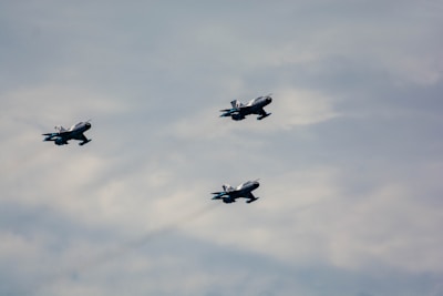 Three military jet aircraft are flying in formation against a cloudy sky. The jets are silver with some markings visible on the tail and wings, and they appear to be moving swiftly through the air.