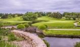 A lush golf course landscape featuring vibrant green grass, manicured lawns, and a variety of trees. A small pond is in the foreground bordered by a stone wall, and there are flowering plants adding color to the scene as well. The overcast sky provides a soft, diffused light over the entire area.