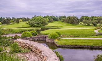 A lush golf course landscape featuring vibrant green grass, manicured lawns, and a variety of trees. A small pond is in the foreground bordered by a stone wall, and there are flowering plants adding color to the scene as well. The overcast sky provides a soft, diffused light over the entire area.