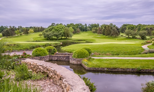 A lush golf course landscape featuring vibrant green grass, manicured lawns, and a variety of trees. A small pond is in the foreground bordered by a stone wall, and there are flowering plants adding color to the scene as well. The overcast sky provides a soft, diffused light over the entire area.