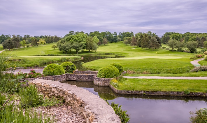 A lush golf course landscape featuring vibrant green grass, manicured lawns, and a variety of trees. A small pond is in the foreground bordered by a stone wall, and there are flowering plants adding color to the scene as well. The overcast sky provides a soft, diffused light over the entire area.