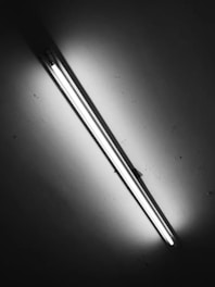 A black and white image of a fluorescent light tube mounted on a ceiling, emitting bright light and casting shadows against the surface.