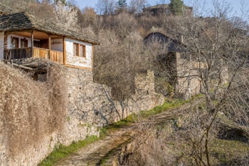 A rustic stone house with a wooden balcony nestled in a hilly, rural landscape. The path leading up to the house is lined with dry vegetation and stone walls, while bare trees are scattered around the area. In the background, more stone structures are visible, blending into the earthy tones of the landscape.