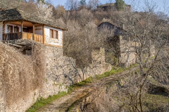 A rustic stone house with a wooden balcony nestled in a hilly, rural landscape. The path leading up to the house is lined with dry vegetation and stone walls, while bare trees are scattered around the area. In the background, more stone structures are visible, blending into the earthy tones of the landscape.