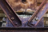 Close-up view of a rusty metal structure with two beams connected by bolts. The surface displays significant corrosion, with visible textures and varying shades of brown and orange. The background is blurred, drawing focus to the metal surface and its weathered condition.