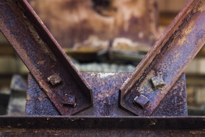 Close-up view of a rusty metal structure with two beams connected by bolts. The surface displays significant corrosion, with visible textures and varying shades of brown and orange. The background is blurred, drawing focus to the metal surface and its weathered condition.
