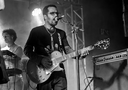 A musician stands on stage playing an electric guitar and singing into a microphone, with another performer in the background playing a keyboard. The atmosphere is enhanced by stage lighting and electronic equipment.