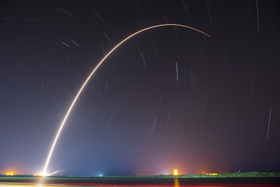 A long exposure photograph captures the launch of a rocket, creating an arc of light against the night sky. Star trails and city lights are visible in the background over a dark landscape.