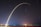A long exposure photograph captures the launch of a rocket, creating an arc of light against the night sky. Star trails and city lights are visible in the background over a dark landscape.