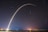 A long exposure photograph captures the launch of a rocket, creating an arc of light against the night sky. Star trails and city lights are visible in the background over a dark landscape.