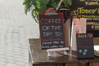 A small café display with a chalkboard sign offering 'Coffee of the Day' for 30. Additional items include a special carrot cake offer with coffee for 60. Nearby, stacks of branded disposable cups are visible beside a container likely for holding coffee. Another chalkboard advertises fresh juice options such as fresh orange juice, raspberry, and pear.