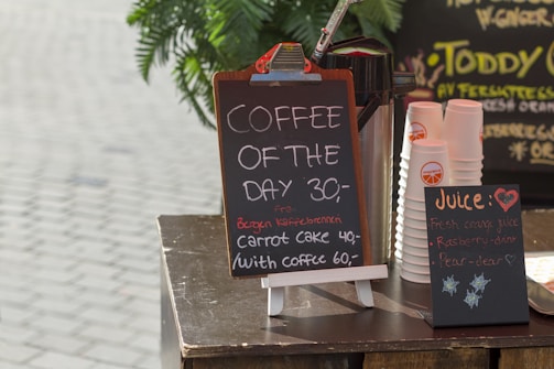 A small café display with a chalkboard sign offering 'Coffee of the Day' for 30. Additional items include a special carrot cake offer with coffee for 60. Nearby, stacks of branded disposable cups are visible beside a container likely for holding coffee. Another chalkboard advertises fresh juice options such as fresh orange juice, raspberry, and pear.