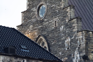 A detailed view of the stone facade of a historic building, featuring a round window and several smaller rectangular openings. The weathered stone exterior displays a variety of textures and colors, complemented by a steeply angled slate roof in the foreground.